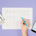 Hand marking days on a calendar with glucose monitor and smartphone app, emphasizing diabetes management.