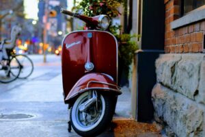 A classic red Vespa scooter parked beside a brick wall in an urban street setting.
