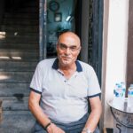 Elderly man sitting outdoors by stairs, smiling warmly at a café entrance.