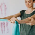 Crop anonymous orthopedist in wristwatch and uniform helping young woman in casual wear reaching arms with elastic tape in doctor office