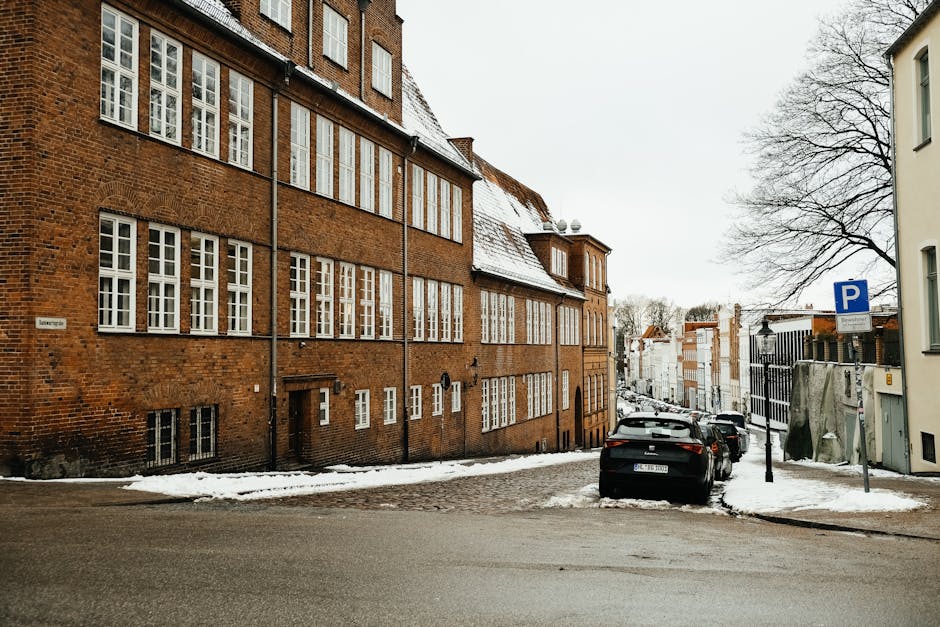 A quiet winter scene of a historic brick building along a snowy urban street with parked cars.