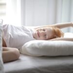 Little adorable child with red hair lying in bed with white linen while trying waking up early morning stretching hands after sleep in light cozy bedroom at home