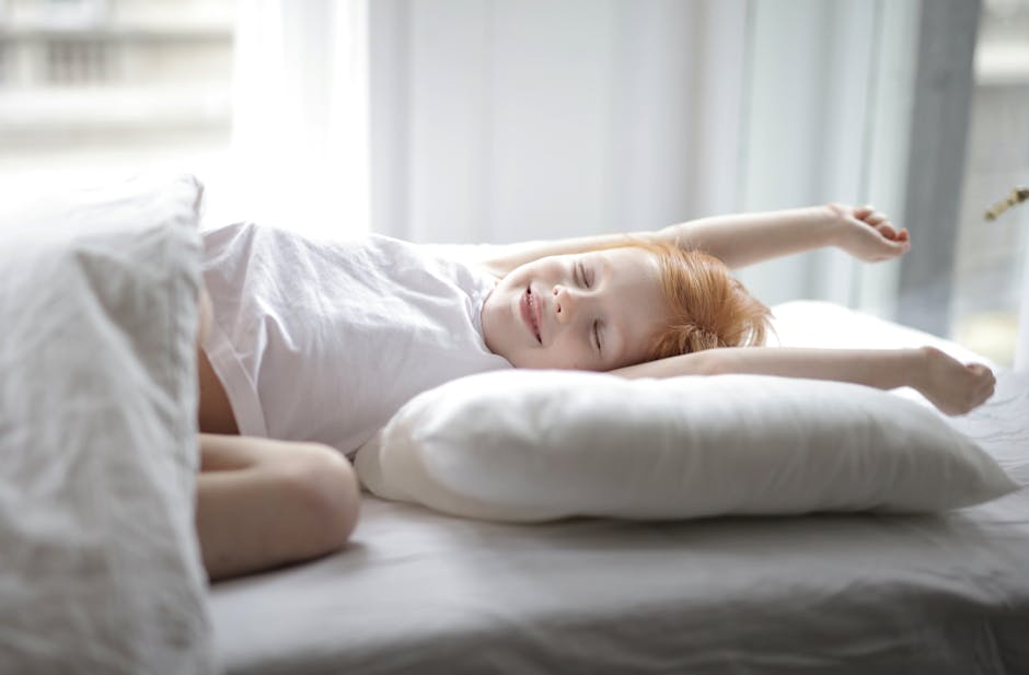 Little adorable child with red hair lying in bed with white linen while trying waking up early morning stretching hands after sleep in light cozy bedroom at home