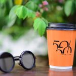 Vibrant orange coffee cup with logo and trendy sunglasses on a wooden table outdoors.