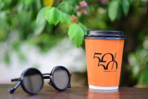 Vibrant orange coffee cup with logo and trendy sunglasses on a wooden table outdoors.