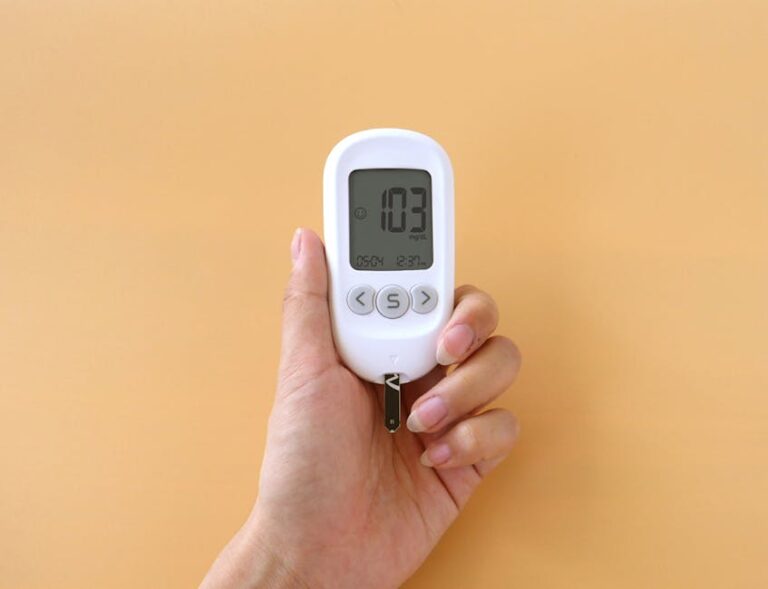 A hand holding a blood glucose meter displaying 103 mg/dL on a neutral background.