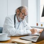 Experienced senior doctor writes a prescription online in a bright, modern office.