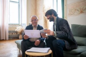An elderly man and a younger man in a casual office setting, discussing documents with smiles.