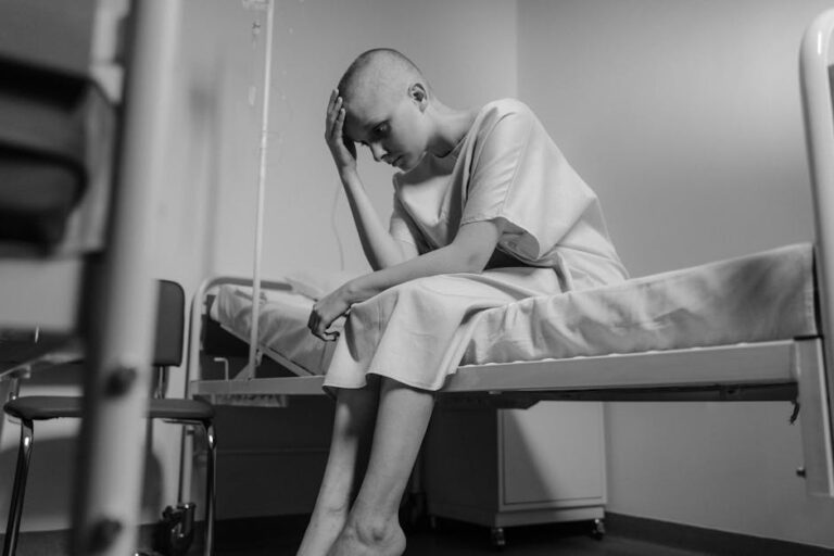Black and white image of a bald woman in a hospital gown sitting thoughtfully on a bed.