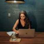 Focused woman writing notes while working on a laptop in a dimly lit room.