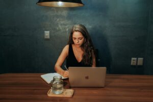 Focused woman writing notes while working on a laptop in a dimly lit room.