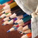 A collection of vibrant colored pencils spilling from a burlap bag on a wooden surface.