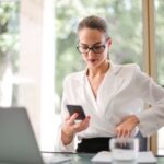 Confident businesswoman in white blazer using smartphone at an office desk with laptop.