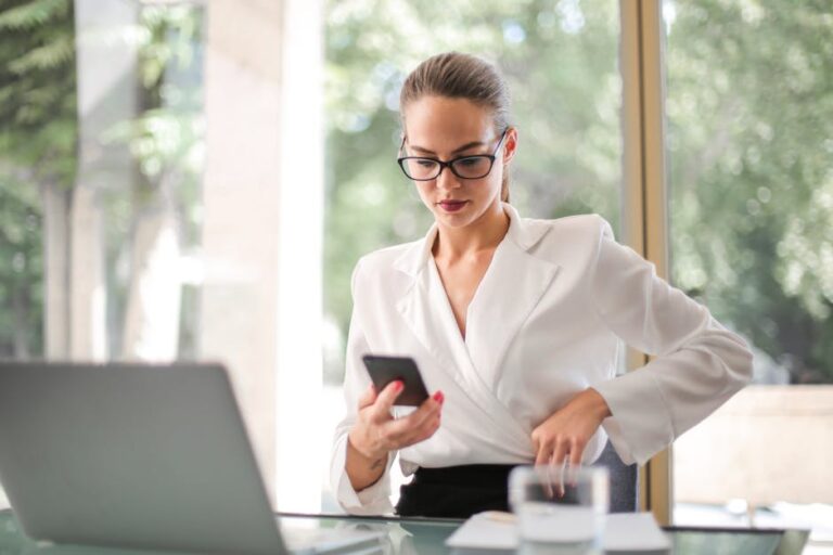 Confident businesswoman in white blazer using smartphone at an office desk with laptop.