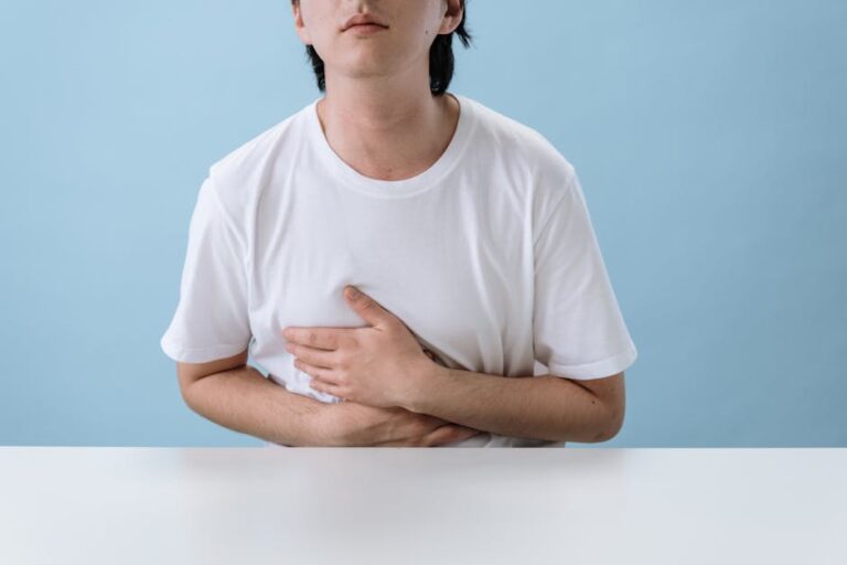 An adult man in a white shirt holds his stomach, indicating pain and discomfort, with a light blue background.