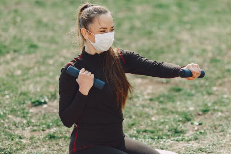 Slim female athlete in sports clothes and sterile mask using dumbbells during workout on grass and looking away in daylight during COVID 19 pandemic