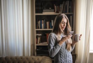Brunette woman in grey sweater texting on smartphone while enjoying coffee indoors by window.