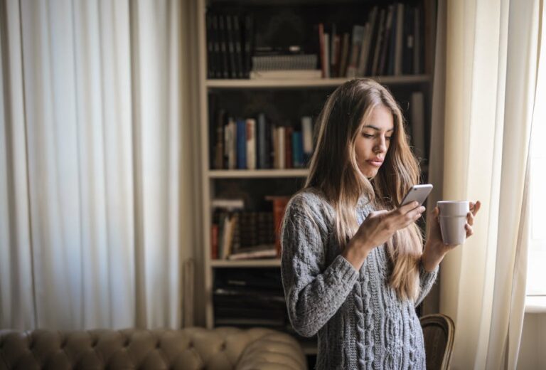 Brunette woman in grey sweater texting on smartphone while enjoying coffee indoors by window.